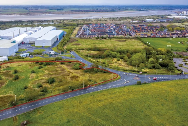 An aerial view highlighting a plot of land with a red line boundary. The area includes overgrown green space bordered by roads, situated between large white industrial warehouses on the left and a residential housing development on the right. The red line clearly outlines the designated plot of land. A river or coastline is visible in the distant background.