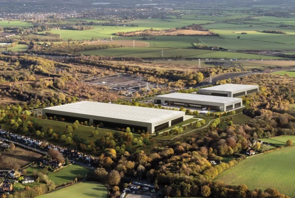 An aerial view of Parkside, a large, industrial development surrounded by green fields and patches of wooded areas. The development consists of multiple modern, warehouse-style buildings with light-colored roofs. The buildings are spaced apart, with landscaped areas featuring trees and greenery between them. To the left, there is a residential neighborhood with rows of houses lining the edge of the wooded area. In the distance, more rural landscapes are visible, with small clusters of houses and industrial sites scattered across the countryside. Power lines and a railway line can also be seen cutting across the fields.